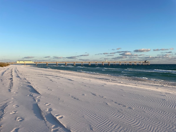 Ein Strand mit einem Steg, der sich ins Wasser erstreckt, sandiger Uferbereich mit Fußabdrücken und einem klaren Himmel.