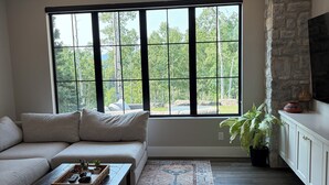 A living room with a sectional sofa, a coffee table, a large window with a view of trees, a stone wall, and a flat-screen TV on a cabinet.