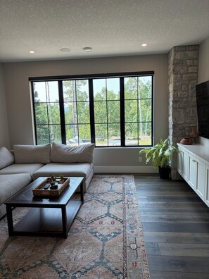 A living room with a sectional sofa, a coffee table, a large window with a view of trees, a stone wall, and a flat-screen TV on a cabinet.