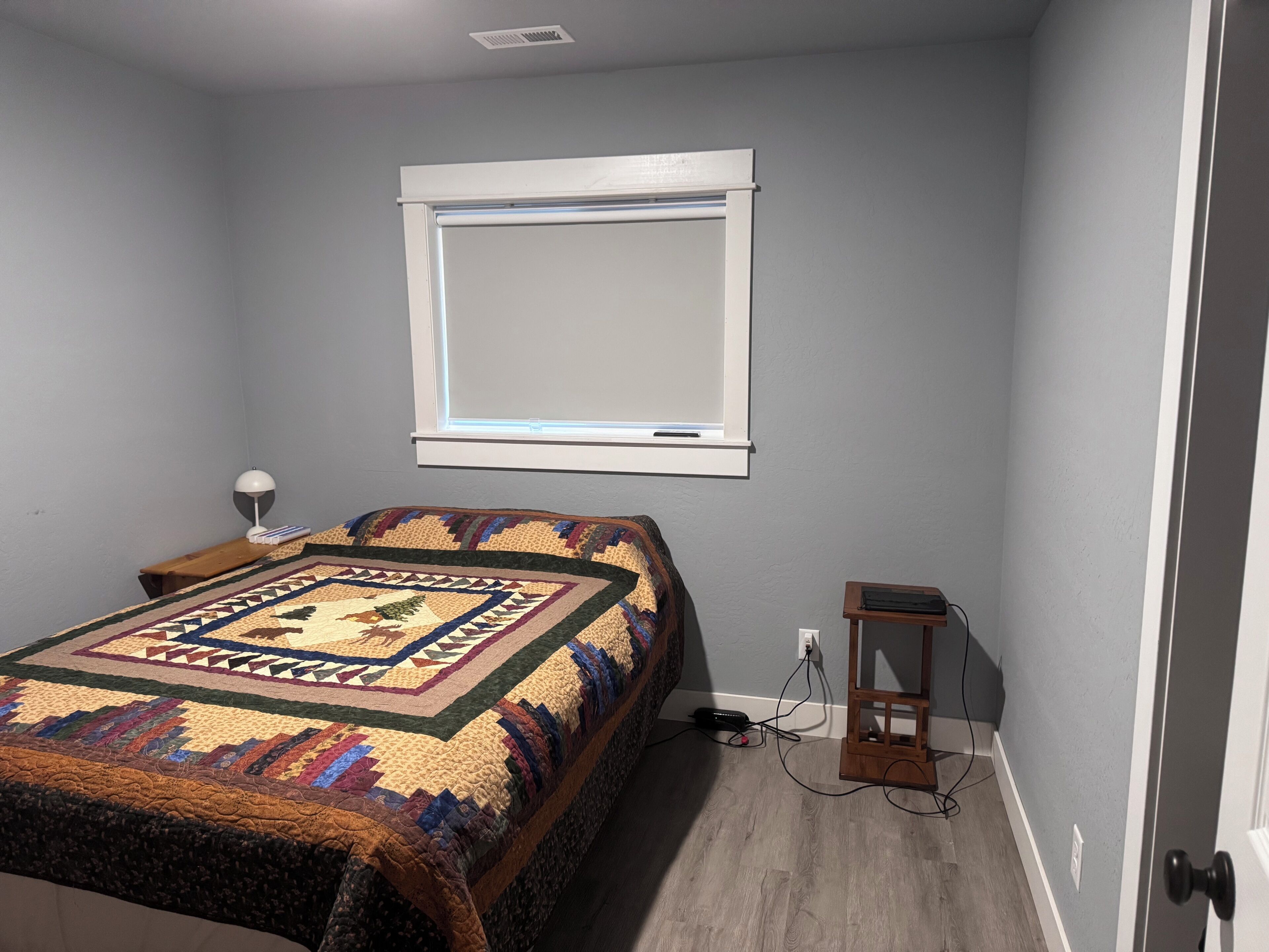 A bedroom with a bed, a nightstand, a lamp, a window, and a wall socket.