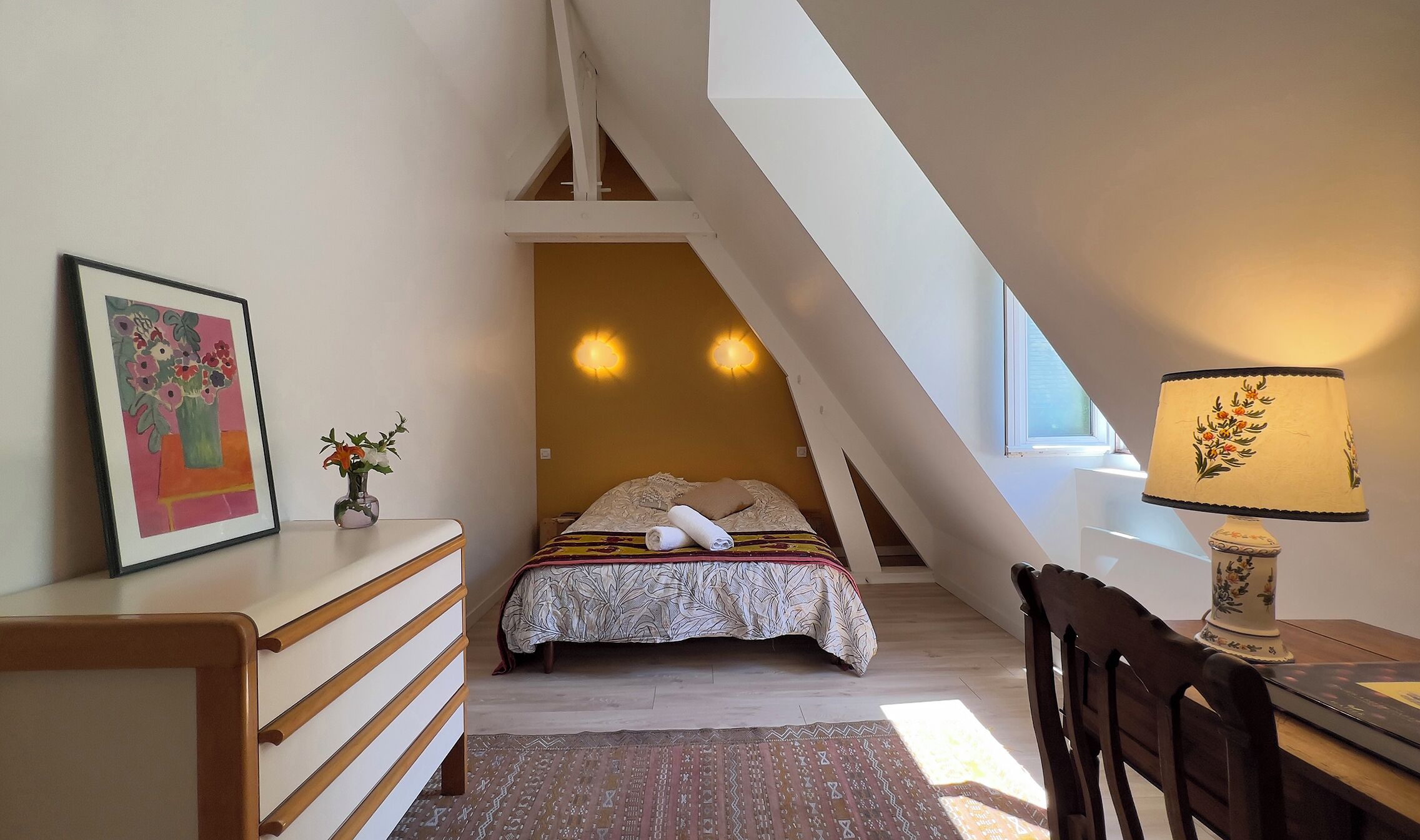 A bedroom with a bed, a desk, a chair, a lamp, and a painting.