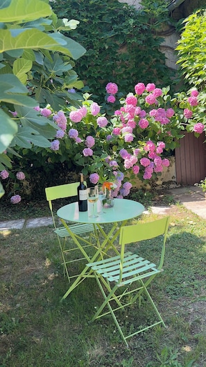 A round table with a bottle and glasses, surrounded by chairs, in a garden with hydrangea bushes.