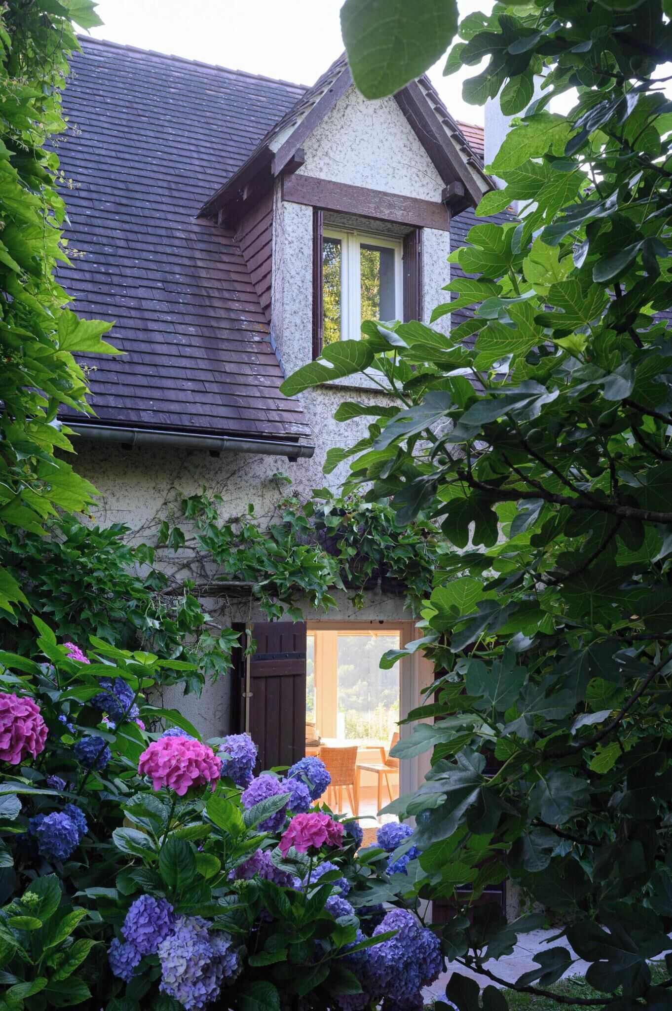 Ein Haus mit geneigtem Dach, einem sichtbaren Fenster und einer Tür. Vor dem Haus befindet sich ein Garten mit Hortensienblüten.