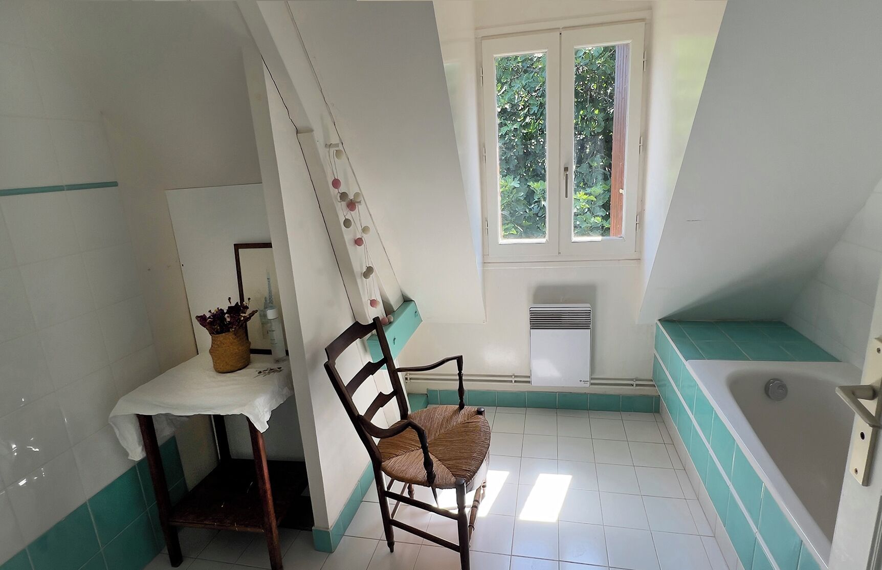 Un baño con una bañera, una silla, una mesita con una planta y una ventana con vista a follaje.
