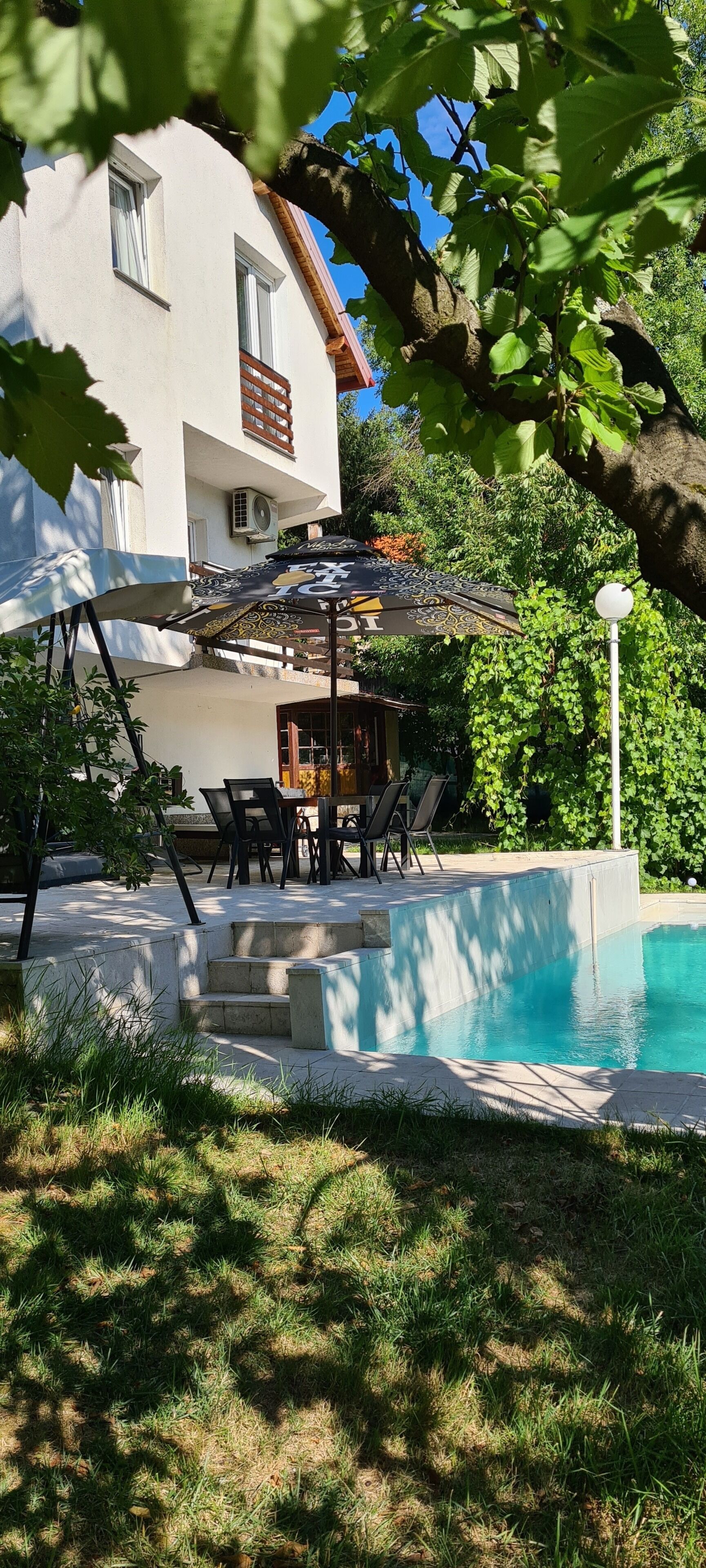 A pool area with a white building, outdoor furniture, and a pergola with an umbrella.
