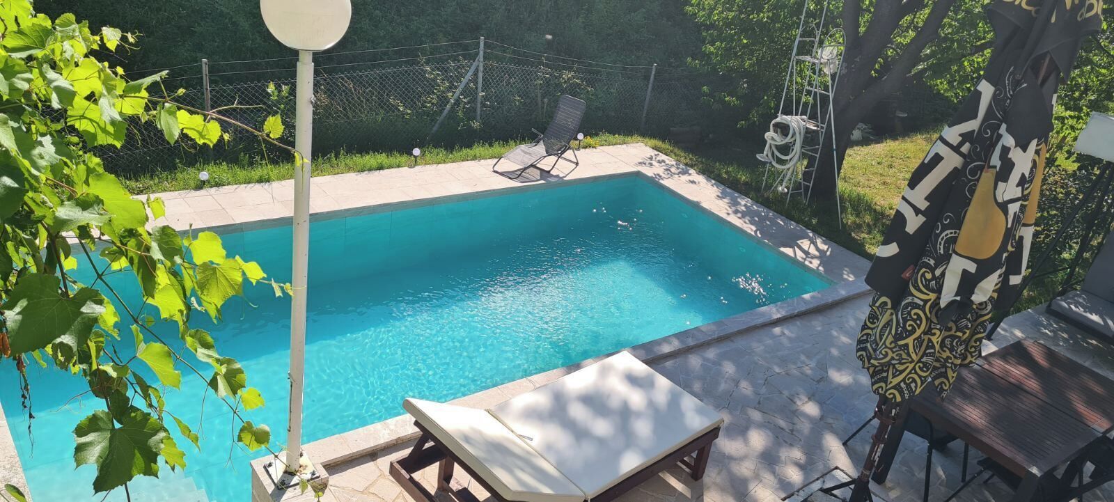 A rectangular swimming pool with a clear blue water surface, surrounded by a stone deck. There are lounge chairs and a table on the deck, and a fence enclosing the area.