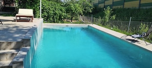 A rectangular swimming pool with a clear blue water surface, surrounded by a concrete deck with steps leading into the pool. There is a lounge chair and a sunshade on the deck.