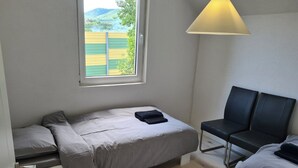 Ein kleines Zimmer mit einem Einzelbett, einem Fenster, einem Nachttisch, einem Stuhl und einer Lampe.