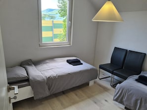 A small, single-bed room with a window, a bedside table, a chair, and a lamp.