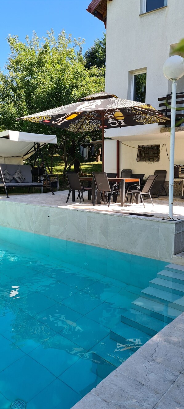 A swimming pool with lounge chairs and umbrellas, a tiled deck, and a building with a window.
