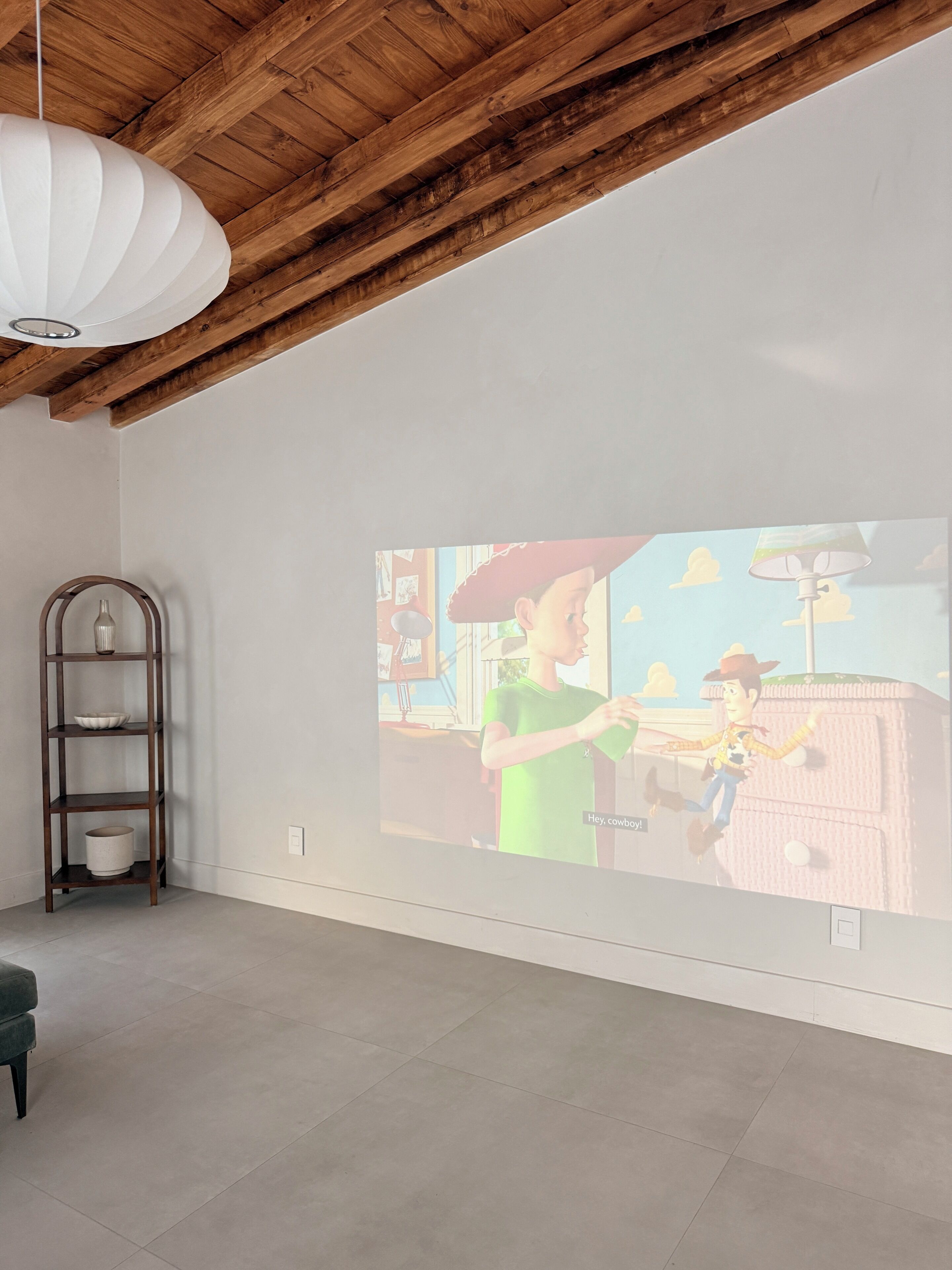 Een kamer met een houten plafond, een witte muur en een projectiescherm waarop een scène met twee personages wordt vertoond.