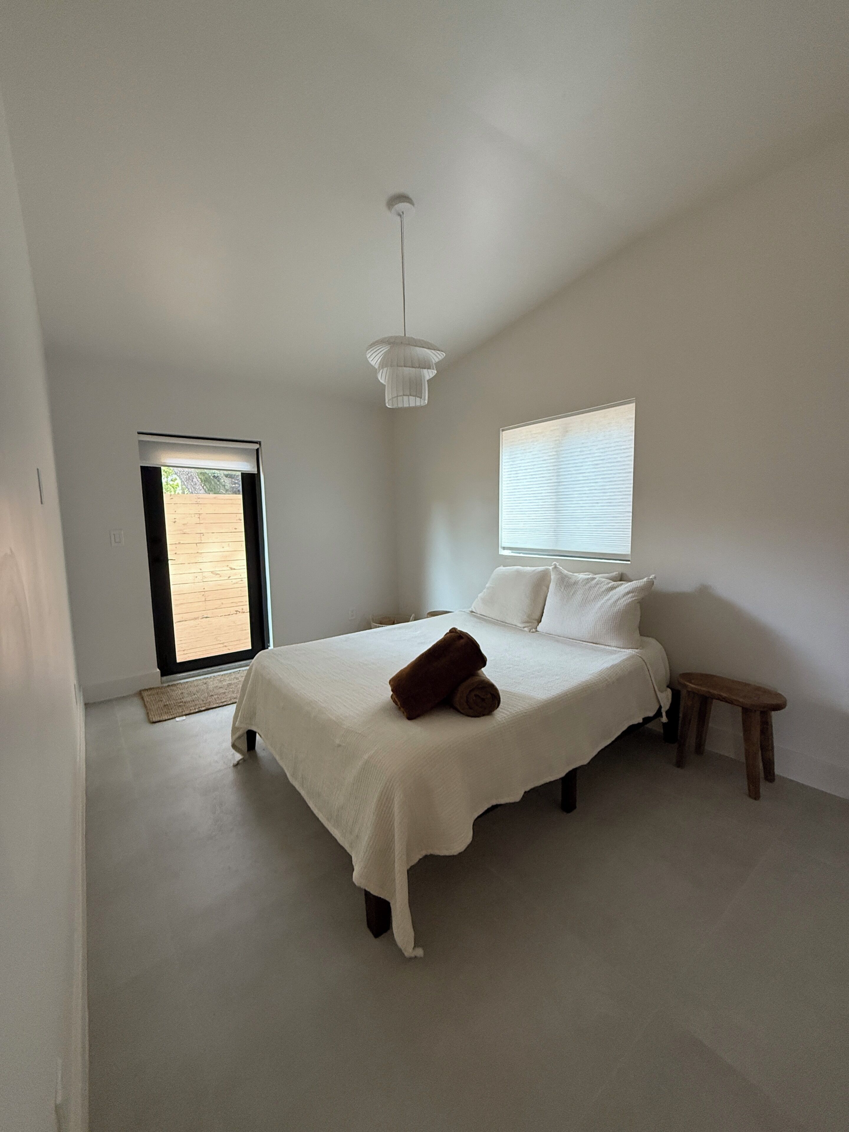 A bedroom with a bed, a small stool, a hanging light fixture, a window with blinds, and a door leading to an outdoor area.
