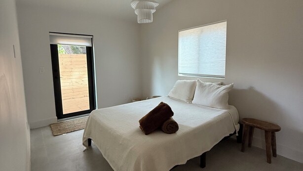 A bedroom with a bed, a small stool, a hanging light fixture, a window with blinds, and a door leading to an outdoor area. - Private Miami home retreat with Oak Tree Deck & Gated Yard / 3 beds, 2 baths (Miami)