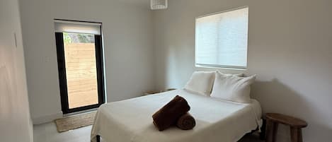 A bedroom with a bed, a small stool, a hanging light fixture, a window with blinds, and a door leading to an outdoor area.