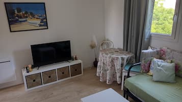 A living room with a flat-screen TV, a dining table with a floral tablecloth, a sofa with cushions, and a painting on the wall.