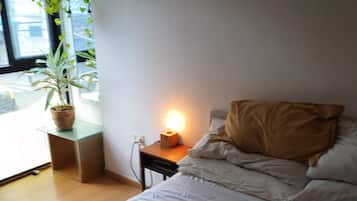 Un dormitorio con cama, mesita de noche, lámpara y una ventana con planta.
