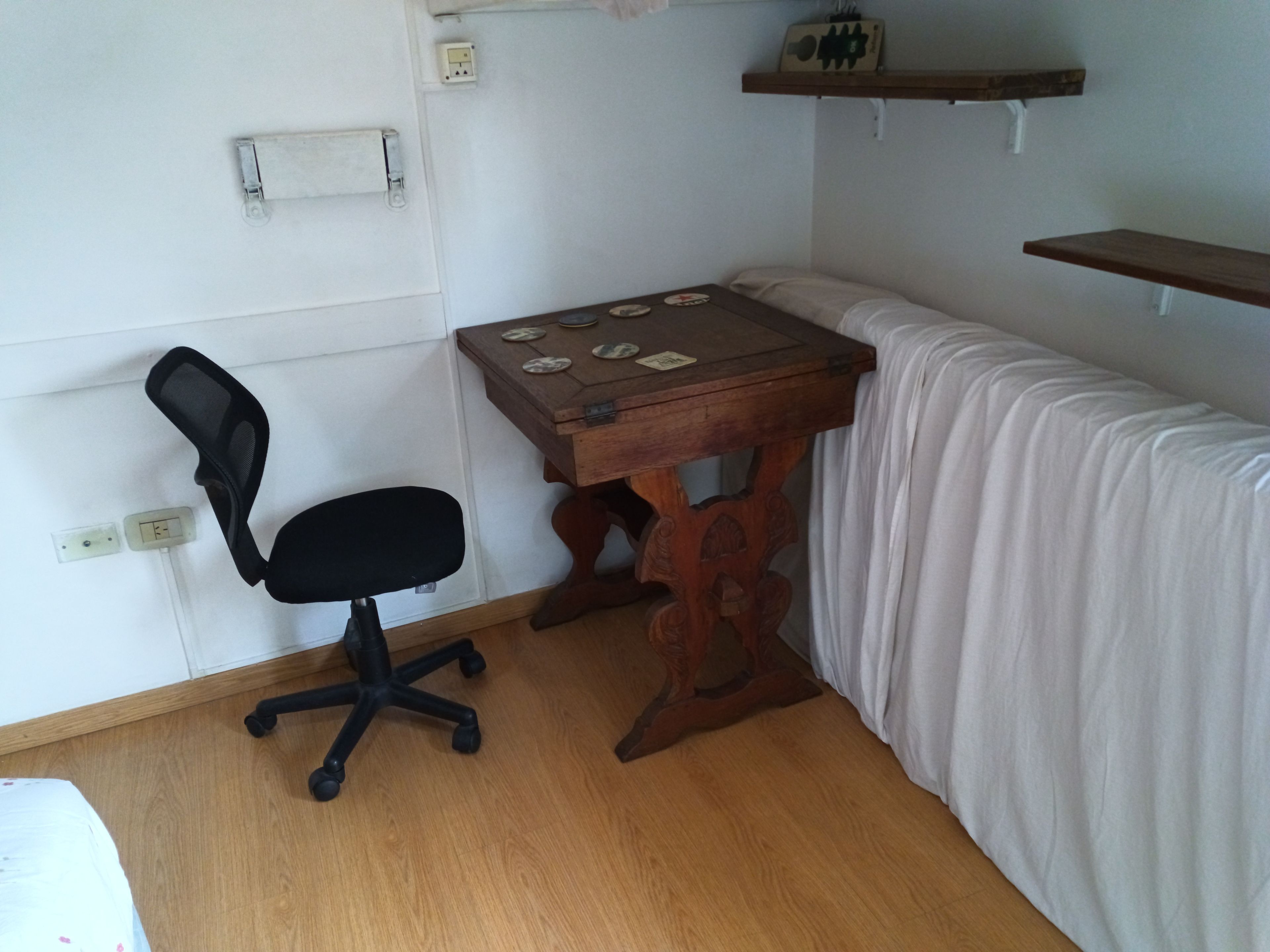 A small room with a wooden desk, a black office chair, a white bed, and a white curtain.