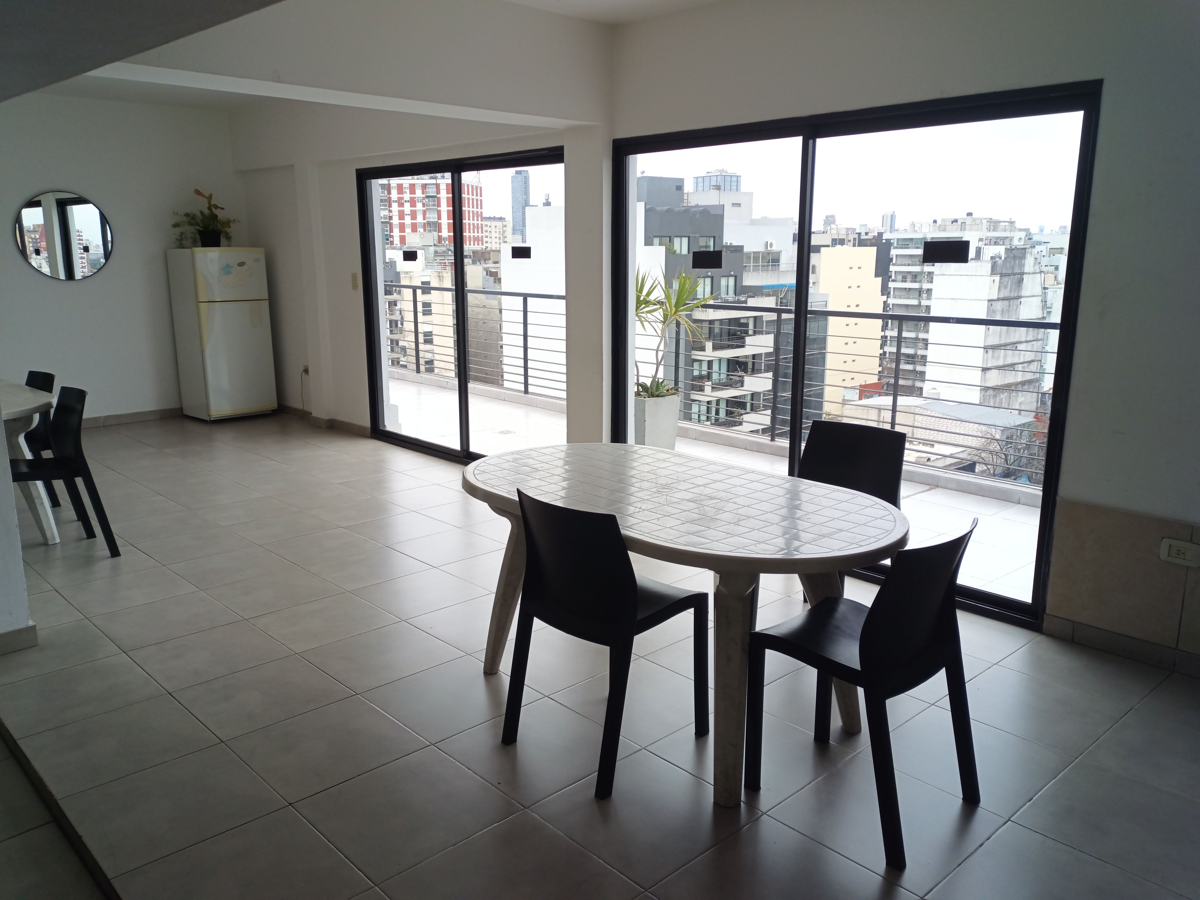A modern room with a round table and chairs, large windows with a city view, a refrigerator, and a potted plant.