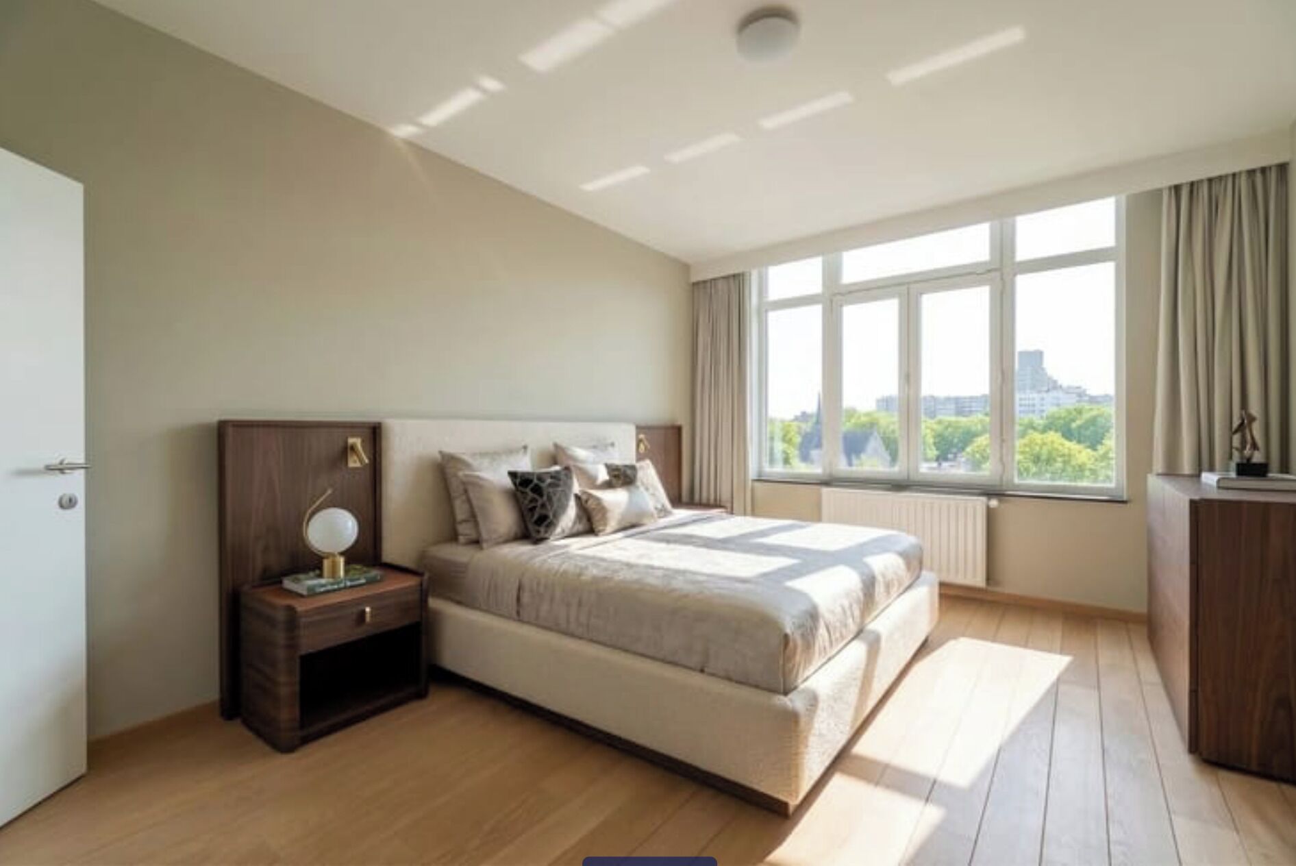 A bedroom with a bed, bedside table, lamp, and a view of the city through large windows.
