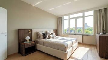 A bedroom with a bed, bedside table, lamp, and a view of the city through large windows.