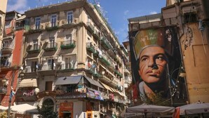 A multi-story building with balconies, a large mural, and various storefronts. - Mia Napoli (Naples)