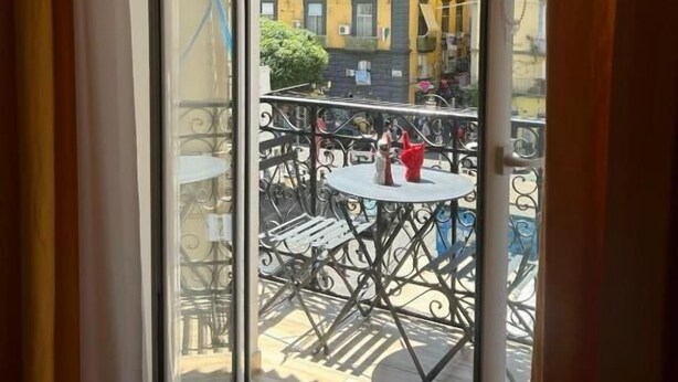 A view through a glass door to an outdoor balcony with a table and chairs, overlooking a yellow building with multiple floors and balconies. - Mia Napoli (Naples)