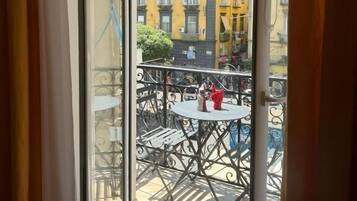 Una vista attraverso una porta vetrata verso un balcone esterno con un tavolo e sedie, affacciato su un edificio giallo con diversi piani e balconi.