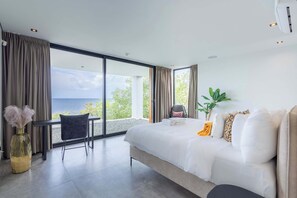 A modern bedroom with a large bed, a seating area with a chair, and a view of the sea. - Villa Coral Breeze | Luxury Villa | Private Beach (Willemstad)