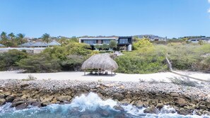 A beachfront property with a thatched-roof structure, a pool, and a view of modern buildings in the background. - Villa Coral Breeze | Luxury Villa | Private Beach (Willemstad)