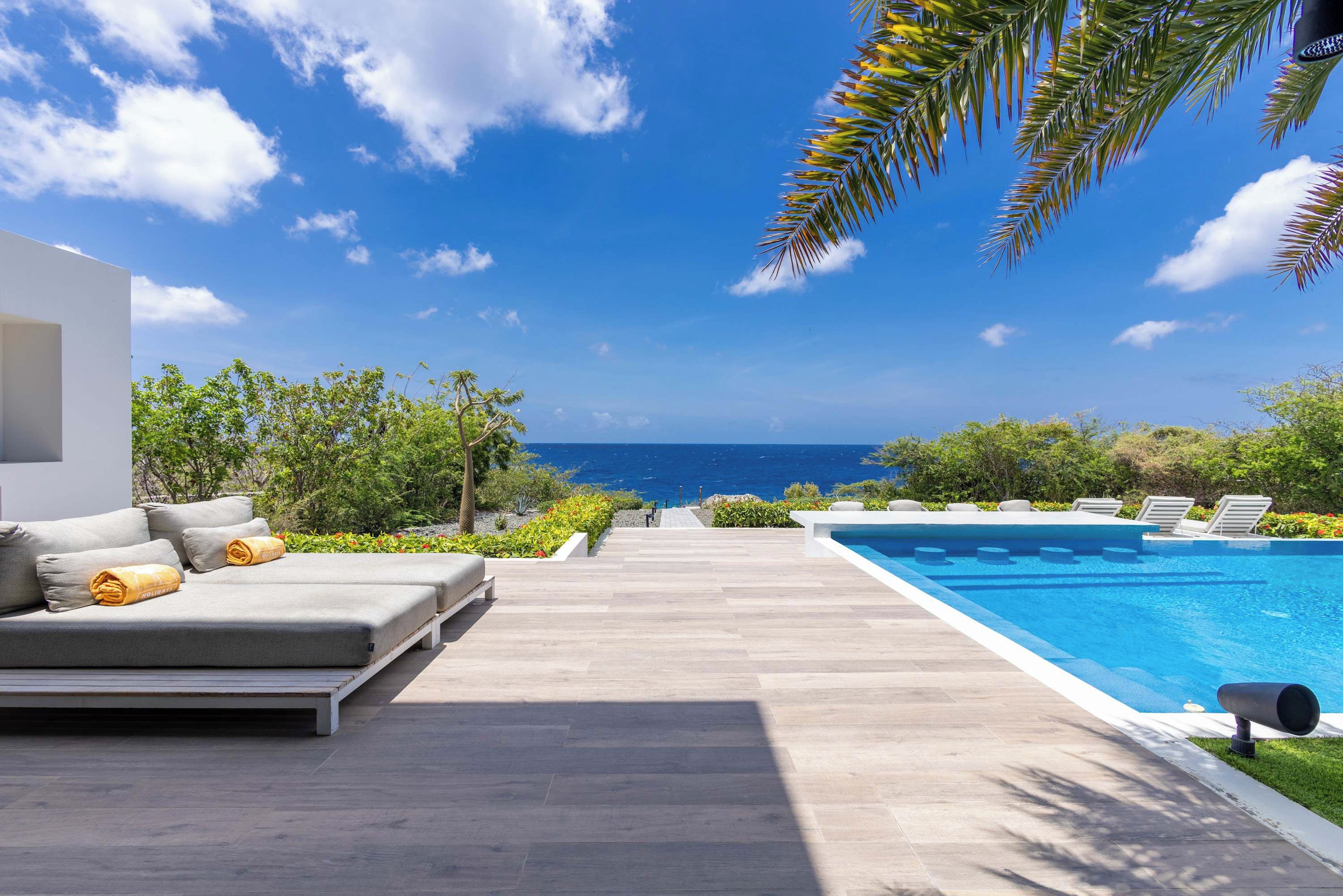A modern outdoor pool area with a clear view of the ocean, a lounge with a sofa and pillows, and a pathway leading to the pool.