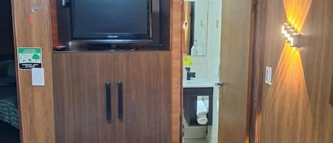 Habitación de hotel con armario de madera, televisor, mesita de noche de vidrio y baño visible a través de una puerta abierta.