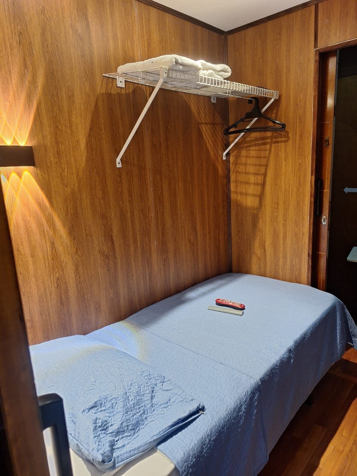 Una habitación pequeña con cama individual, pared de madera, un tendedero blanco y una colcha azul.