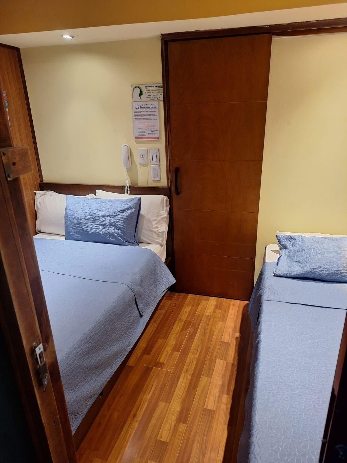 Een hotelkamer met twee bedden, houten vloeren, een deur en een aan de muur bevestigd telefoon.