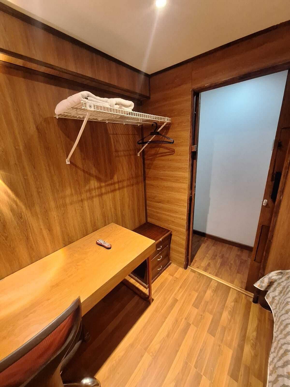 A compact room with a wooden floor, a small desk with drawers, a hanging clothes rack, and a door.