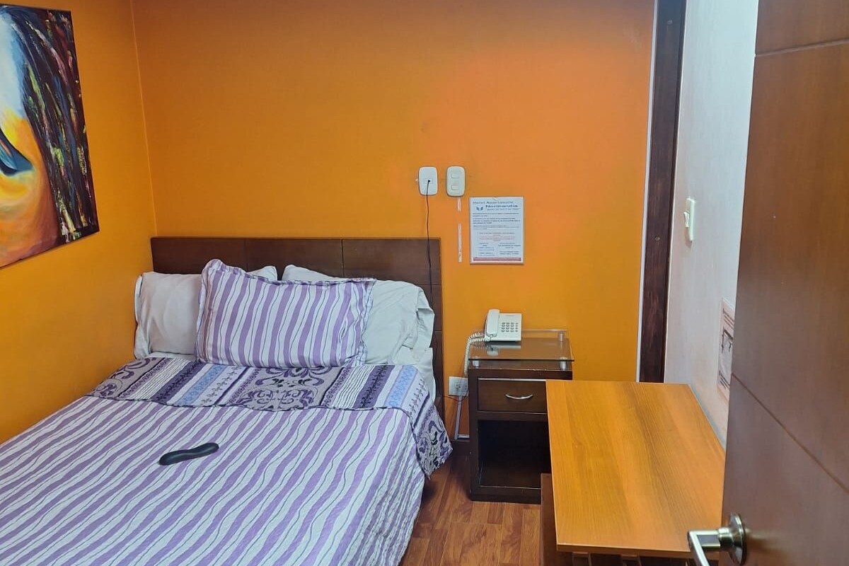 A hotel room with a bed, bedside table, and a small wooden desk.