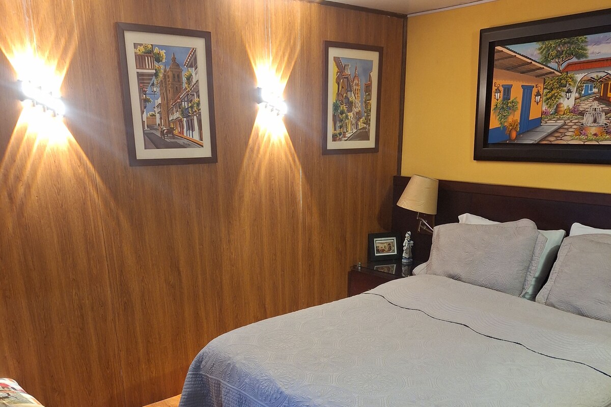 A hotel room with a large bed, a bedside table with a lamp, a chair, and two wall-mounted lights.