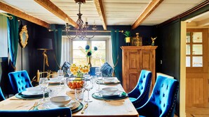 A dining room with a wooden ceiling, a chandelier, a large window with curtains, and a blue upholstered dining table set for four. - Charming Cottage for 4 in Dorset (Brixham)