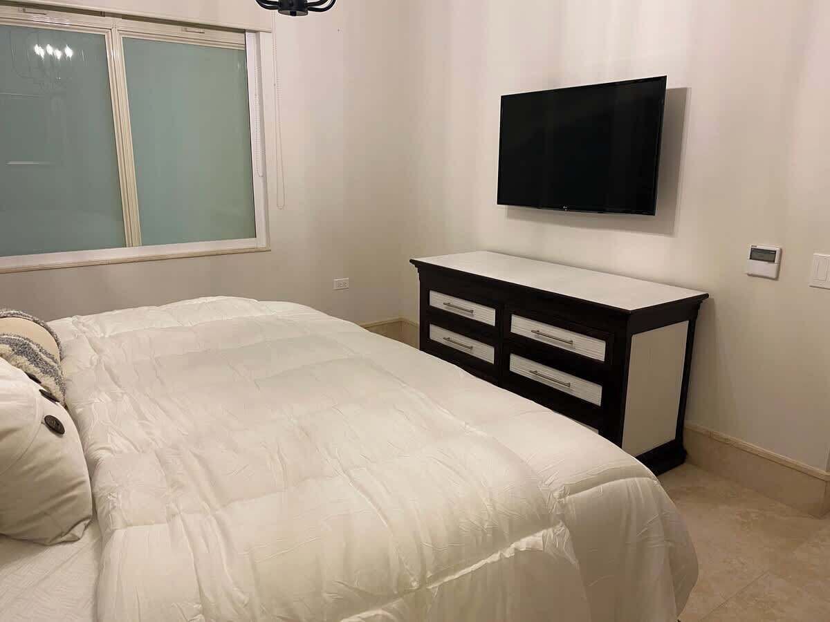 A bedroom with a bed, a bedside table, a TV mounted on the wall, and a window.