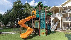 Un parque infantil con un tobogán, estructuras para escalar y un edificio con balcones y escaleras al fondo.