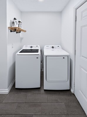 A laundry room with two white front-loading washers and dryers, a wooden shelf with cleaning supplies, and a white door.