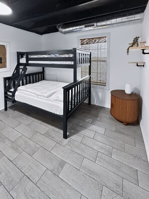 A bunk bed room with a window, a round wooden side table, and a radiator on the ceiling.