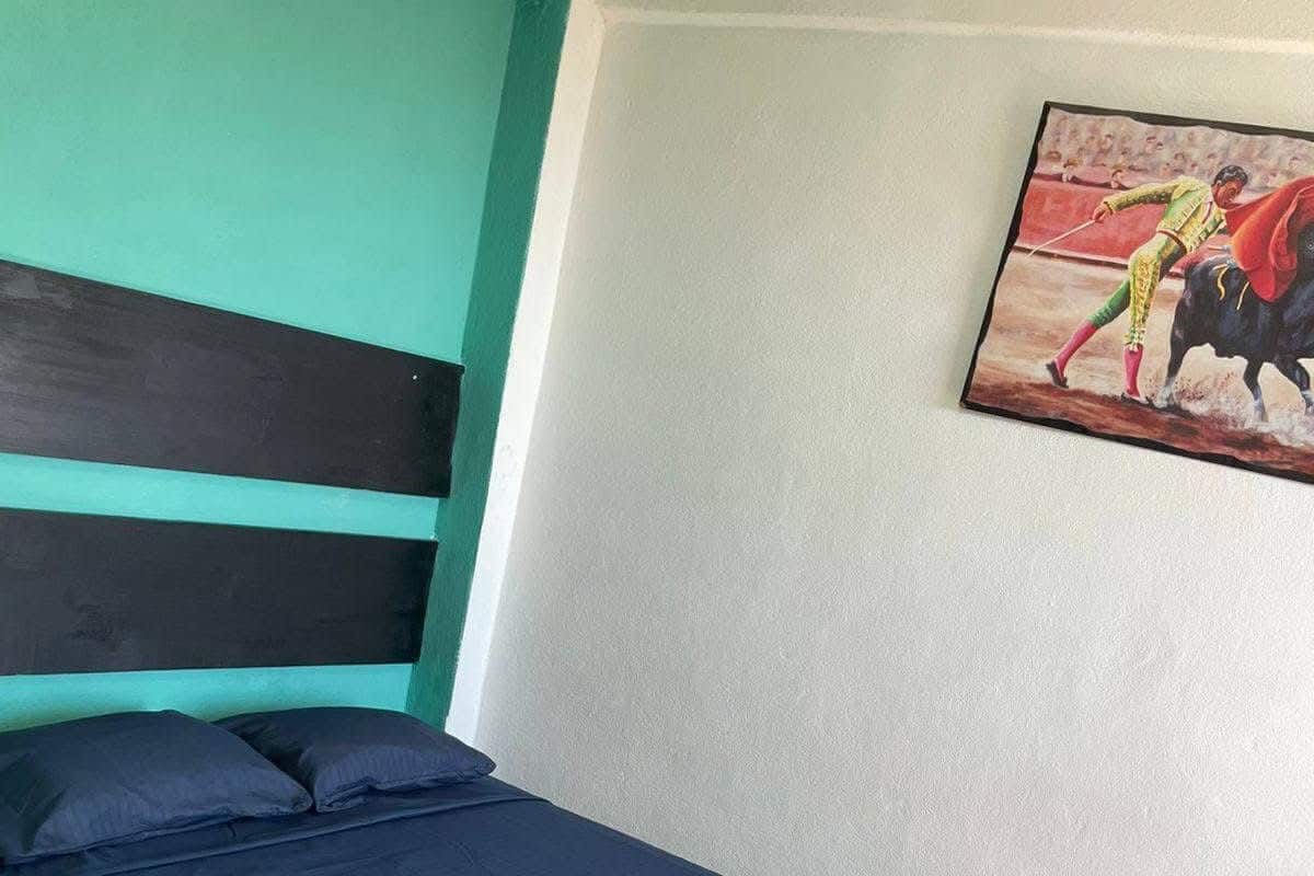 Una cama con una colcha azul y roja, una pared verde azulado y un cuadro de un torero.