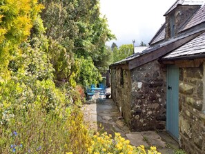 A narrow stone pathway leading to a rustic stone building with a blue door, surrounded by lush greenery and flowering plants. - Cottage in Snowdonia National Park (Dolgellau)