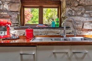 A kitchen with a stone wall, a window with a view of trees, a red compost bin, and a green bottle. - Cottage in Snowdonia National Park (Dolgellau)
