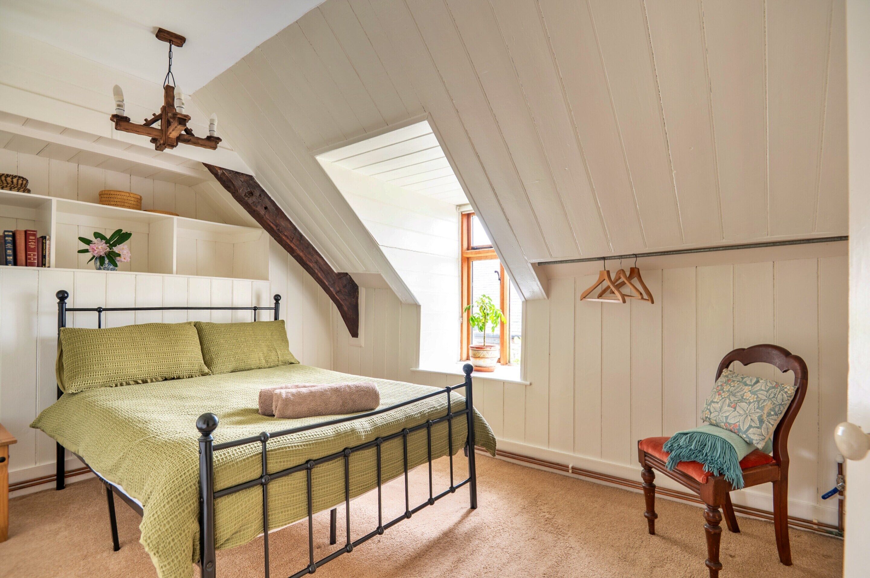 A bedroom with a sloped ceiling, a bed with a green blanket, a wooden chair, a hanging light fixture, and a window with a plant.