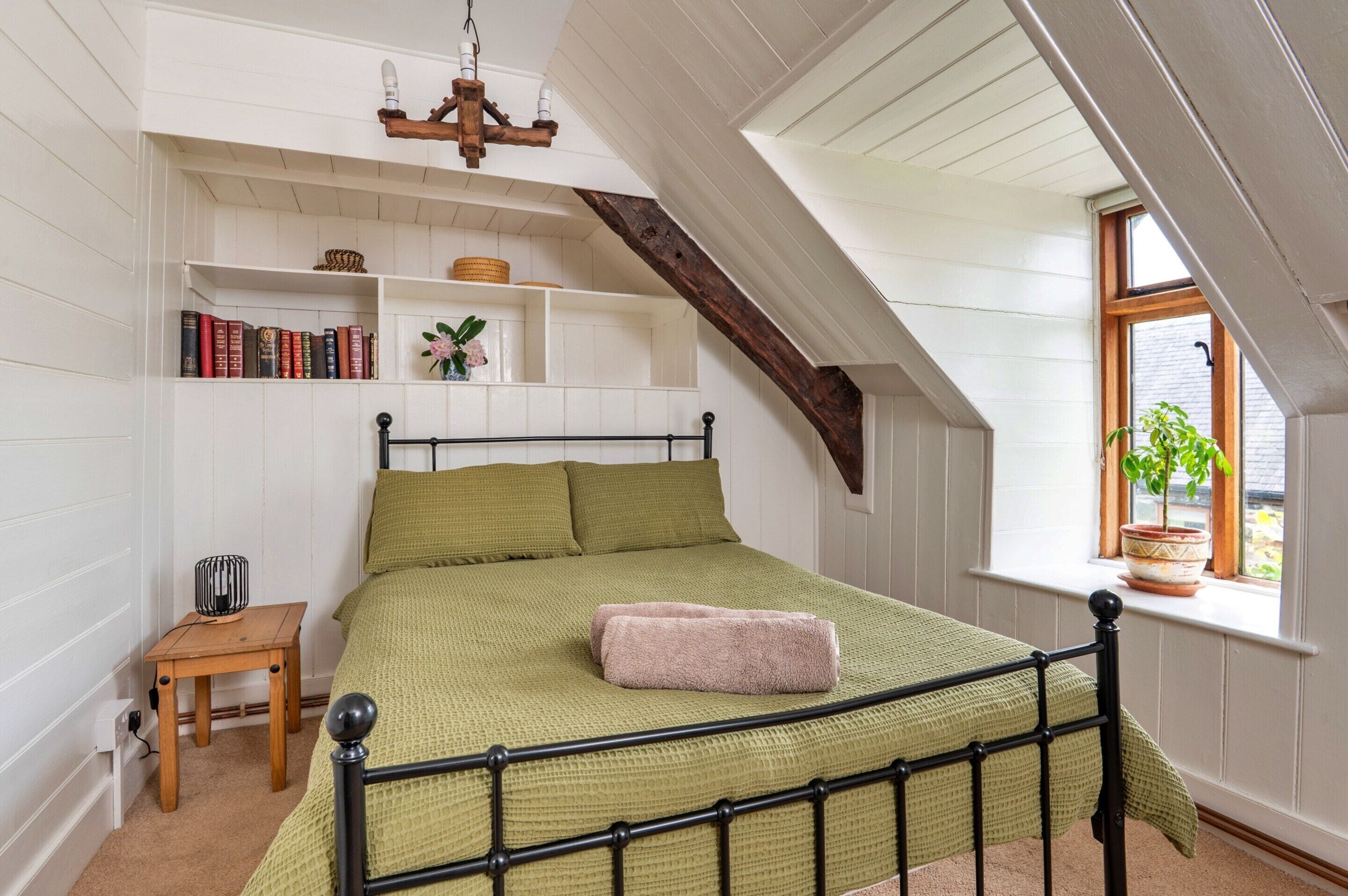 Een slaapkamer met een schuine plafond, een bed met metalen frame, een nachtkastje, een kleine stoel, een boekenplank en een raam met een plant.