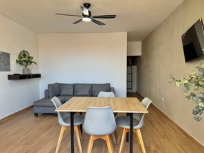 A modern living room with a grey sofa, a wooden dining table with four chairs, a ceiling fan, and a mounted TV. - Vive la Zona Dorada, Boca de Mar by Partner Host (Mazatlán)