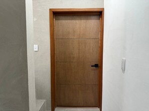 Eine geschlossene Holztür mit schwarzem Griff, weißen Wänden und einem Lichtschalter an der Wand.