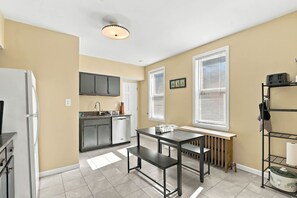 A modern kitchen with a refrigerator, stove, and sink, a dining table with benches, and a radiator under a window. - Walk to E Carson St · 4BR Sleeps 10 · South Side (Pittsburgh)