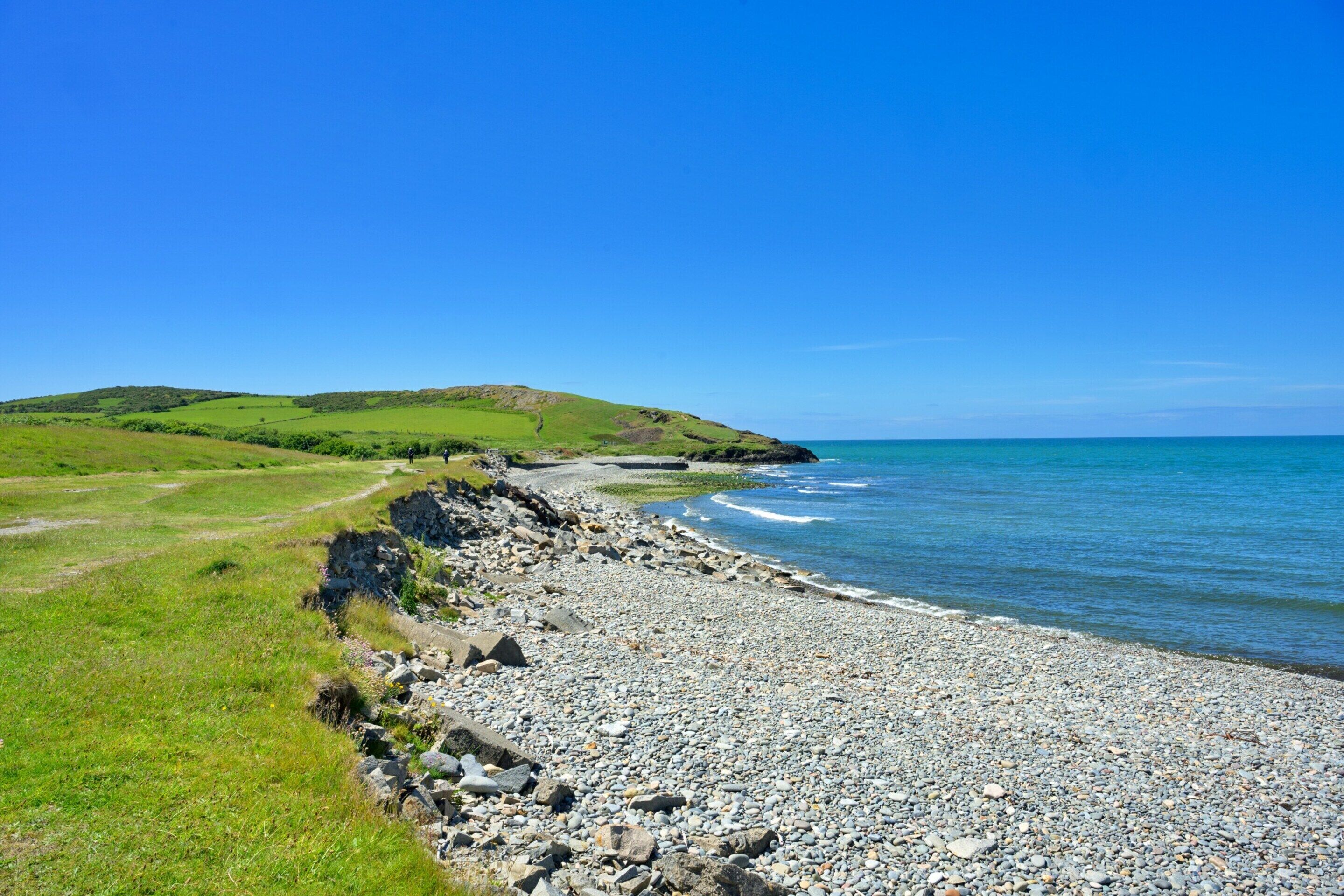 Eine Küstenlandschaft mit kiesigem Strand, grünen Feldern und blauem Himmel.